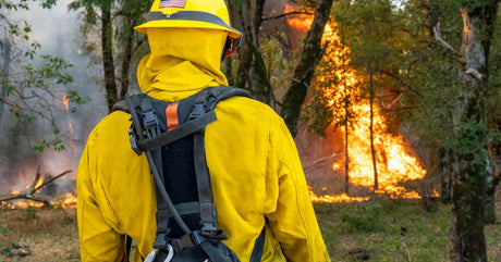 A wildland firefighter wearing bright yellow protective gear and a helmet stands in a forest while watching a fire.