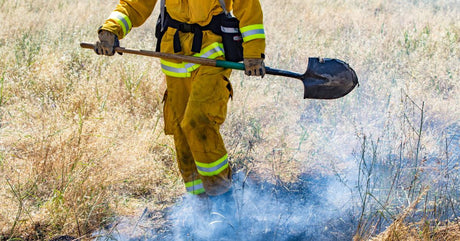 A wildland firefighter in full gear from the chest down, holding a shovel and approaching smoldering grass.