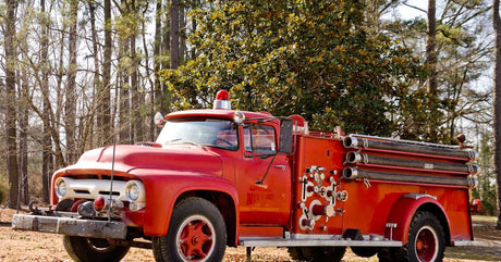 A bright red antique firetruck is parked a dry patch of ground in a forest, backed by a grove of trees.