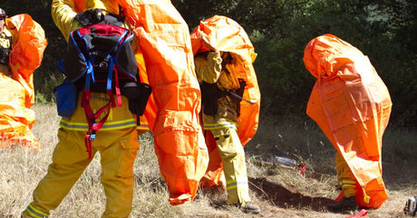 Four wildland firefighters in full gear practice using bright orange portable fire shelters in a grassy clearing.