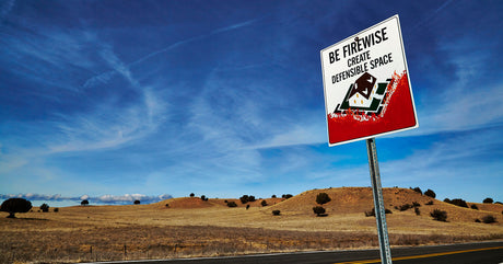 In a scrubby looking area, a road sign with a house surrounded by flames states: 'Be Firewise: Create Defensible Space.'